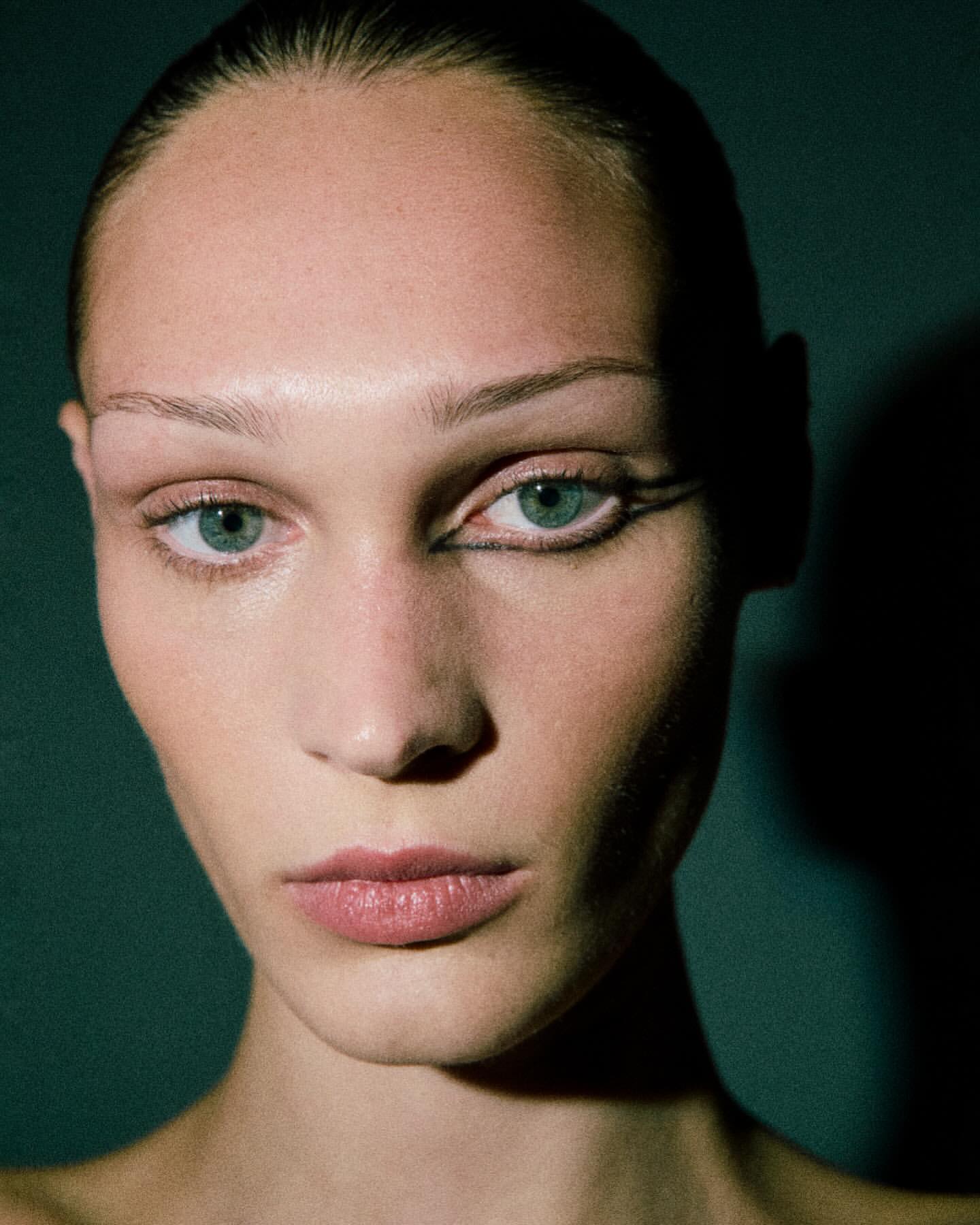 Close-up portrait of a young adult woman with fair skin, light hair, and artistic black eyeliner, set against a dark greenish-grey background.
