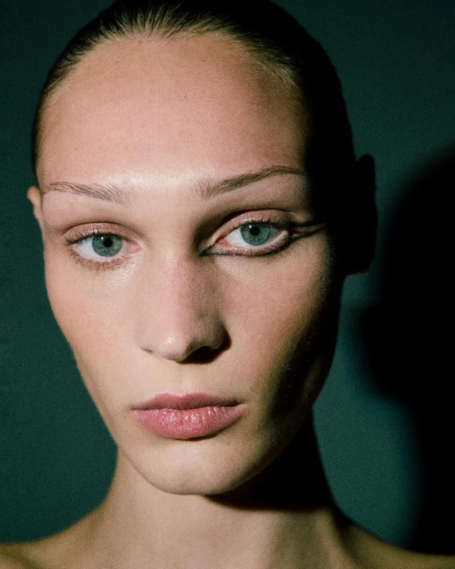 Close-up portrait of a young adult woman with fair skin, light hair, and artistic black eyeliner, set against a dark greenish-grey background.