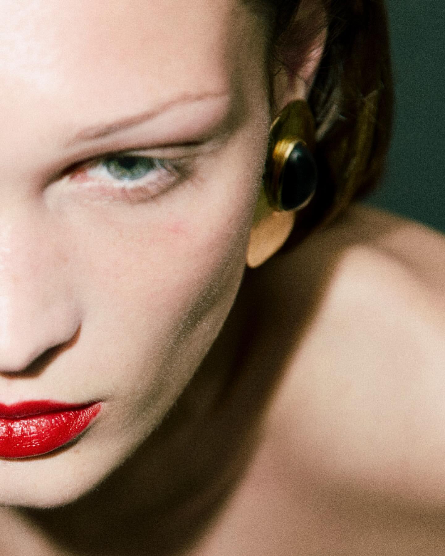 Close-up portrait of a woman with red lipstick, pale skin, and an oversized gold earring with a black stone accent.