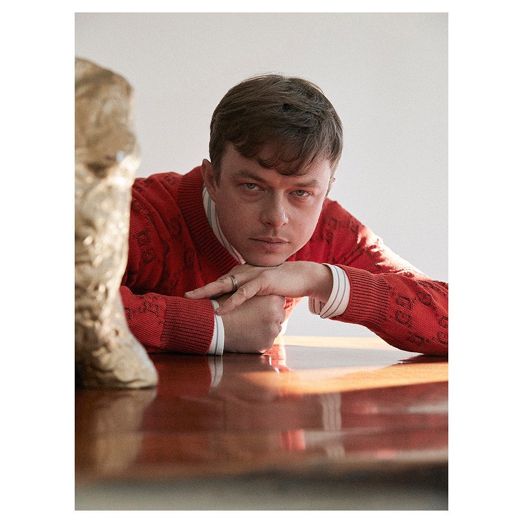 Portrait of a male wearing a red sweater, resting his arms on a glossy table with a sculpture nearby in a brightly lit indoor space.