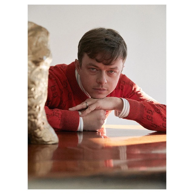 Portrait of a male wearing a red sweater, resting his arms on a glossy table with a sculpture nearby in a brightly lit indoor space.