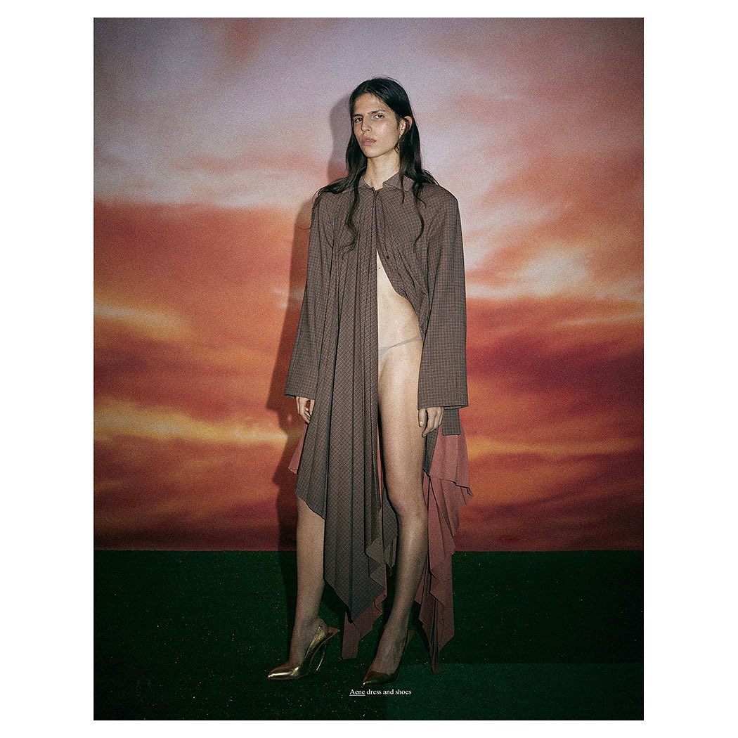Fashion editorial shot of a woman in a long brown jacket and gold heels against a backdrop mimicking a sunset sky.