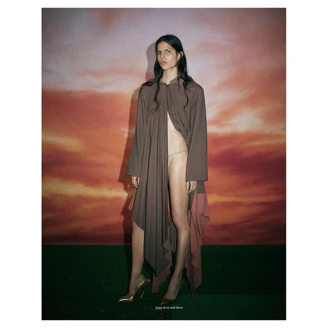 Fashion editorial shot of a woman in a long brown jacket and gold heels against a backdrop mimicking a sunset sky.