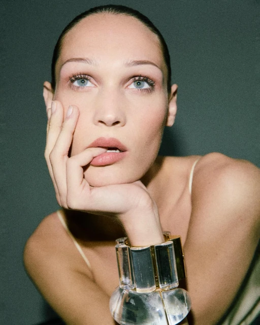 Close-up portrait of a woman with light skin, blue eyes and slicked back hair, wearing a clear bracelet, resting her face on her hand.
