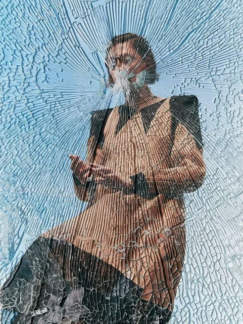 Fashion portrait of a woman in front of a shattered glass window with a blue sky background, wearing an elegant tan dress.