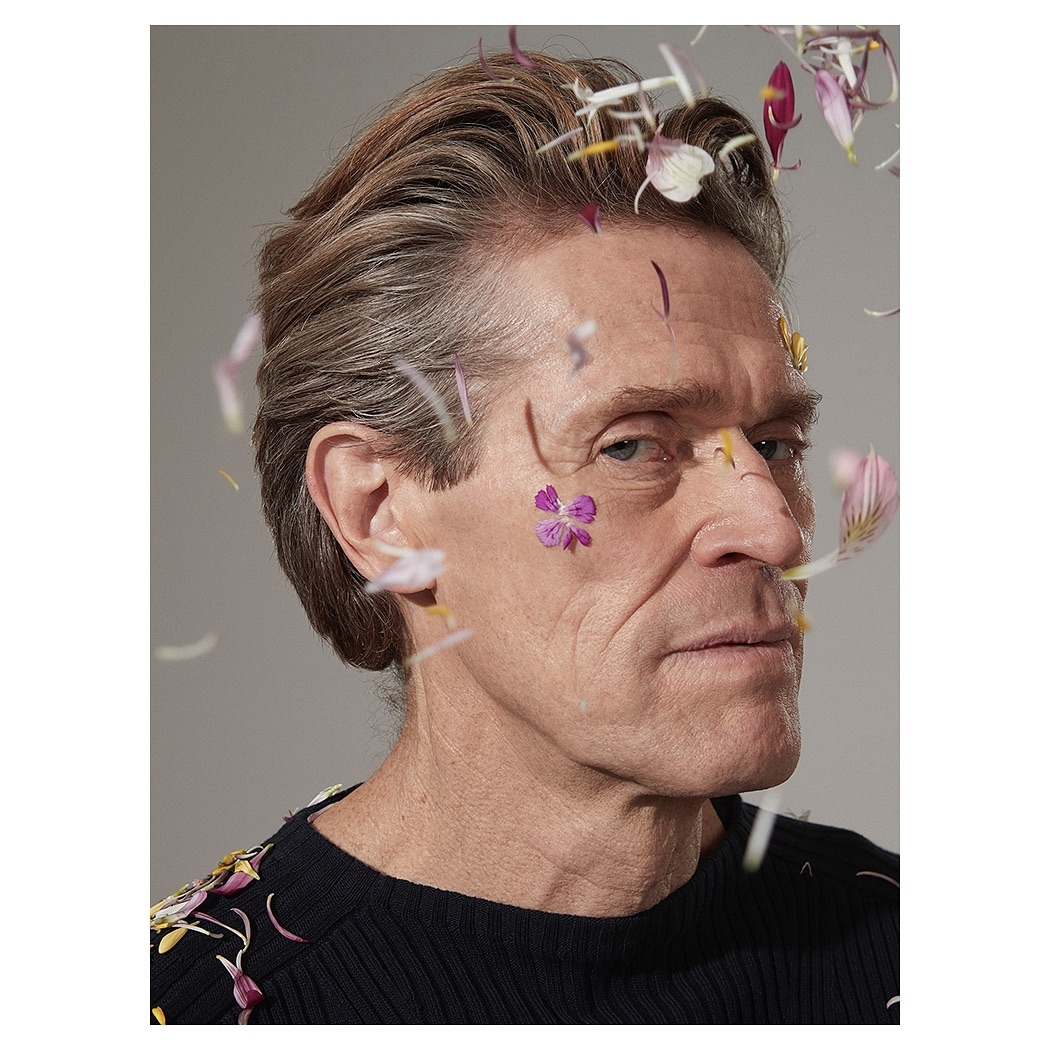 Close-up portrait of Willem Dafoe with flower petals, against a grey backdrop, wearing a black ribbed shirt, soft light.