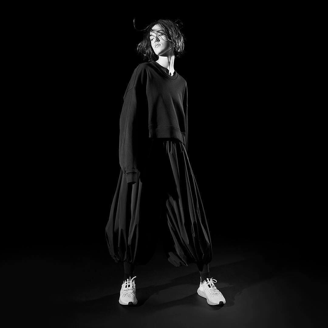 A woman poses in a black top and large, flowing black pants, complemented by white athletic shoes, set against a stark black backdrop.