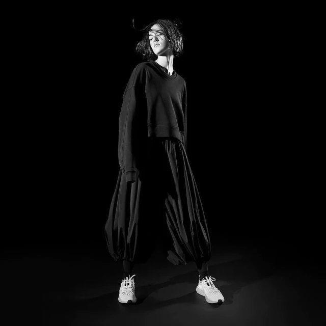 A woman poses in a black top and large, flowing black pants, complemented by white athletic shoes, set against a stark black backdrop.