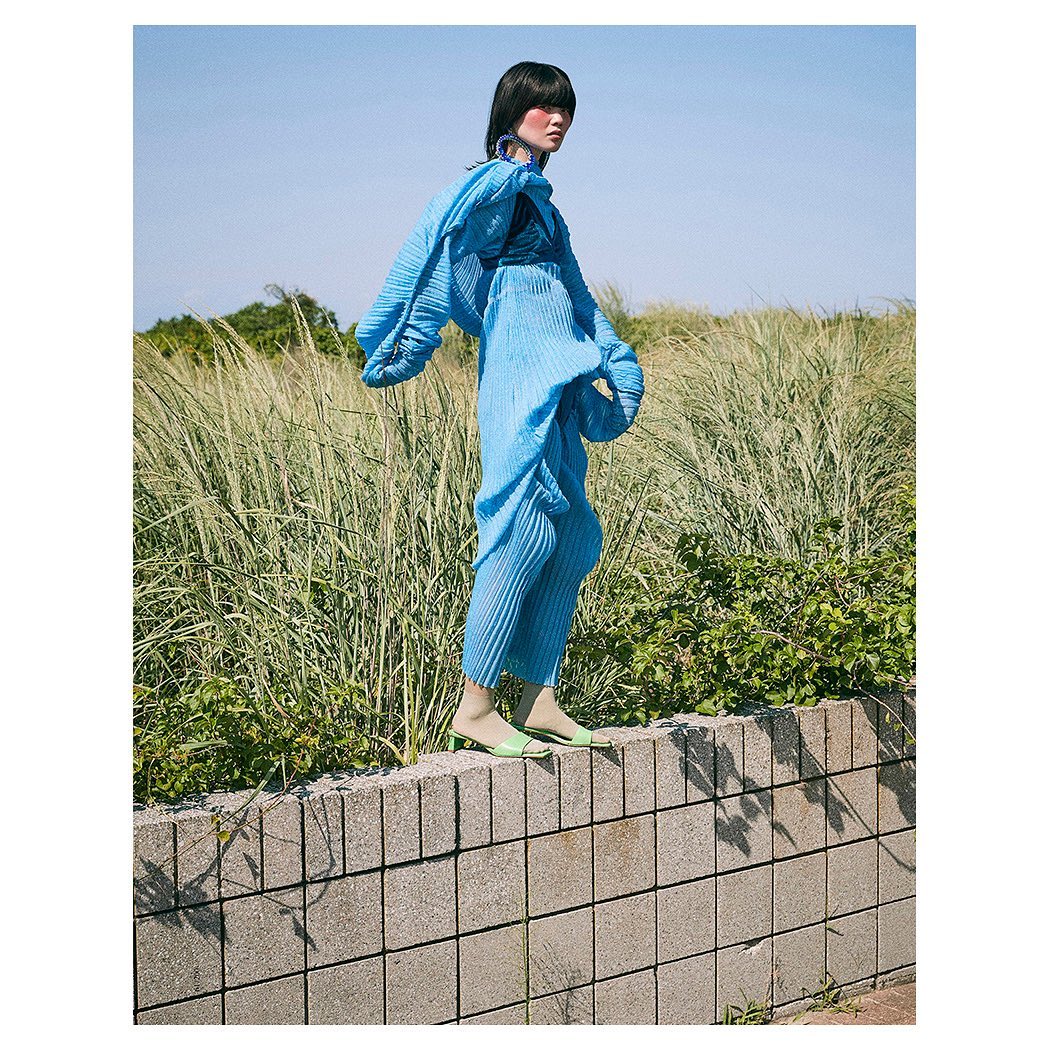 Fashionable woman in a pleated turquoise outfit striking a pose on a wall with tall grass and blue sky creating a natural backdrop.