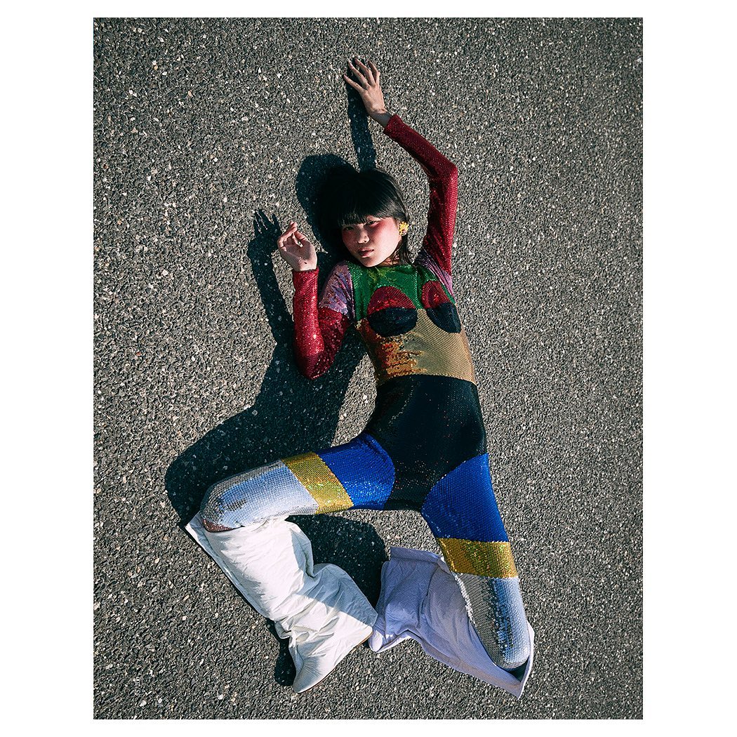 Young woman in a colorful sequined jumpsuit poses on an asphalt surface with white boots, casting a shadow on a sunny day.