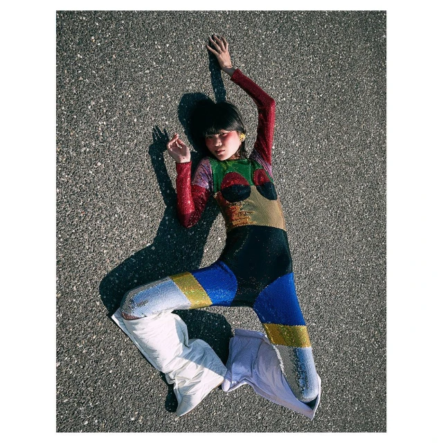 Young woman in a colorful sequined jumpsuit poses on an asphalt surface with white boots, casting a shadow on a sunny day.