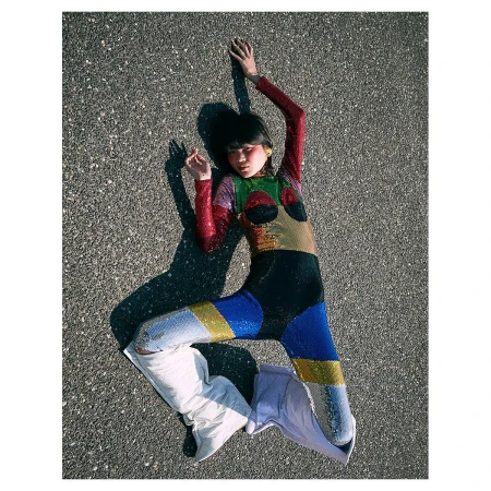 Young woman in a colorful sequined jumpsuit poses on an asphalt surface with white boots, casting a shadow on a sunny day.