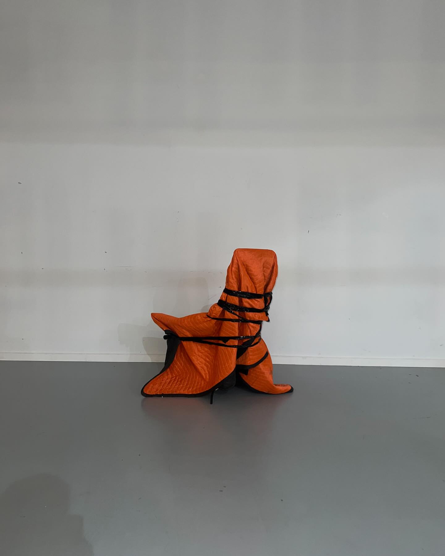Armchair wrapped in orange quilted furniture pad and black rope, centered against a white wall and grey floor.