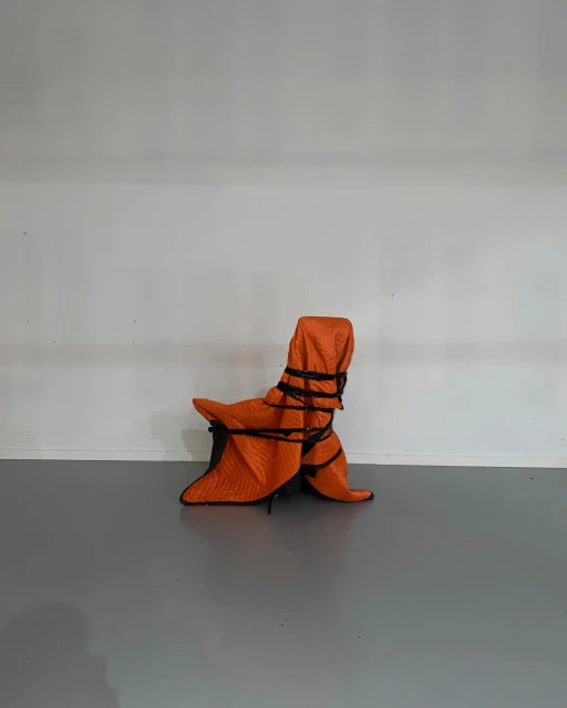 Armchair wrapped in orange quilted furniture pad and black rope, centered against a white wall and grey floor.
