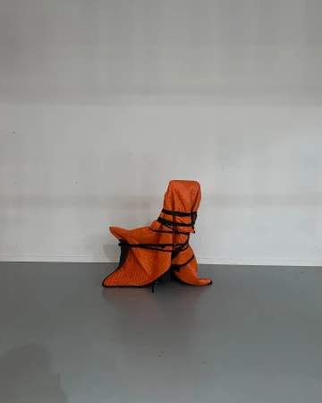 Armchair wrapped in orange quilted furniture pad and black rope, centered against a white wall and grey floor.