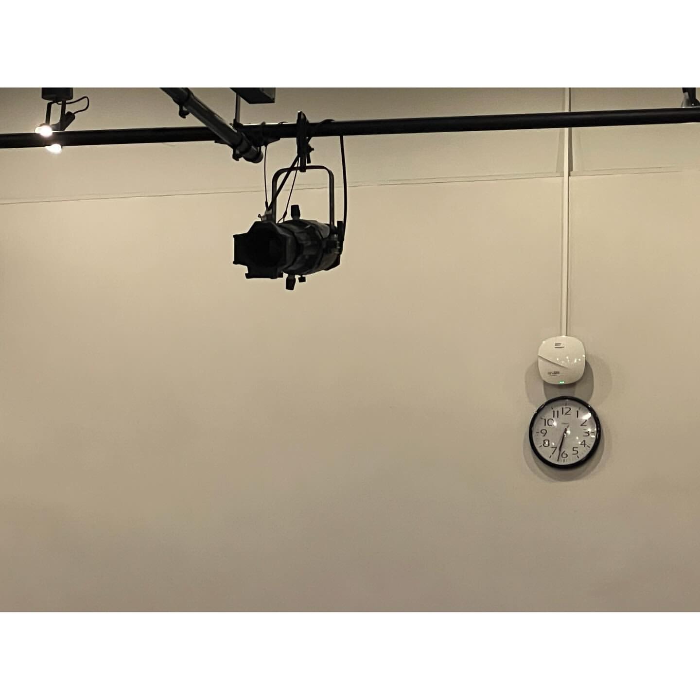 Indoor shot featuring a black stage light hanging from a metal rail on a beige wall, accompanied by a clock and a white sensor.
