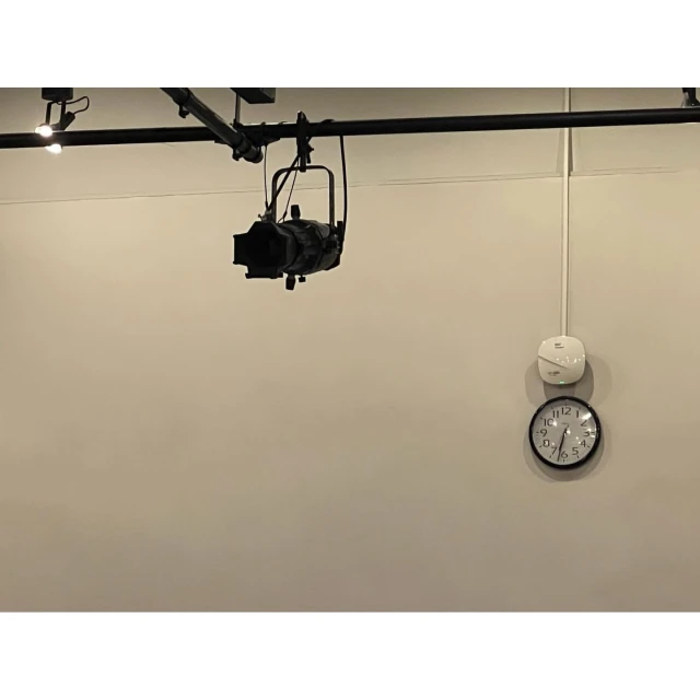 Indoor shot featuring a black stage light hanging from a metal rail on a beige wall, accompanied by a clock and a white sensor.