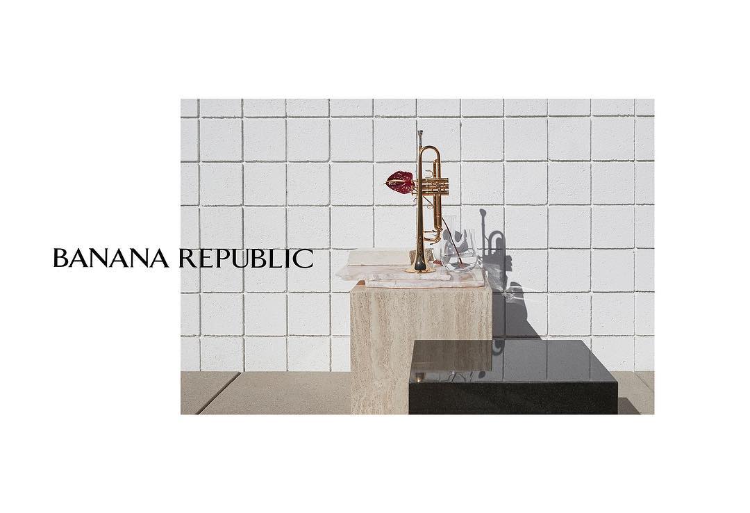 Still life with a gold trumpet adorned with a red flower on a stone pedestal against a textured white brick wall backdrop. 