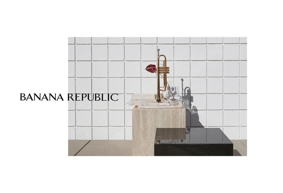 Still life with a gold trumpet adorned with a red flower on a stone pedestal against a textured white brick wall backdrop. 