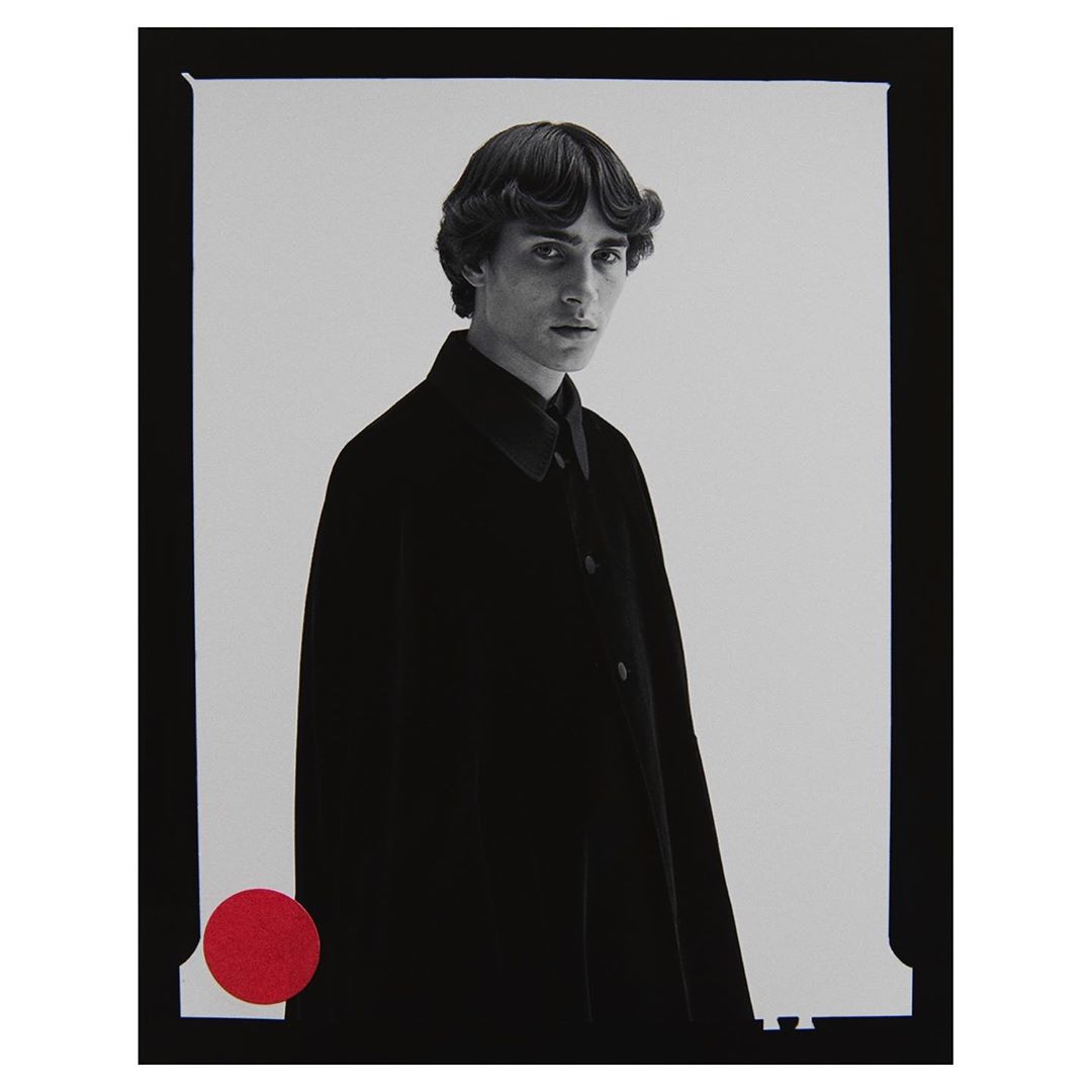 Portrait of a slender adult male model in a black coat, with dark hair and a red circle detail on a monochrome background in a studio.