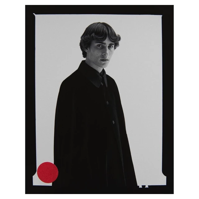 Portrait of a slender adult male model in a black coat, with dark hair and a red circle detail on a monochrome background in a studio.