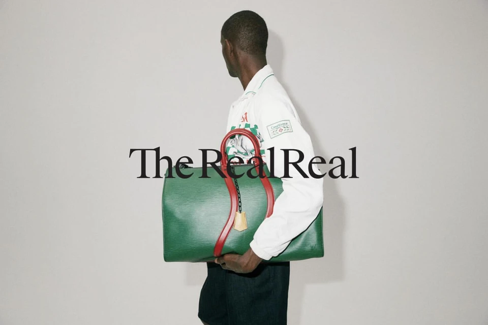 A person carries a green leather duffel bag with red straps in a studio shot with simple background, featuring the 'The RealReal' logo.