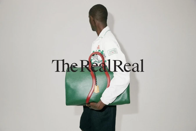 A person carries a green leather duffel bag with red straps in a studio shot with simple background, featuring the 'The RealReal' logo.