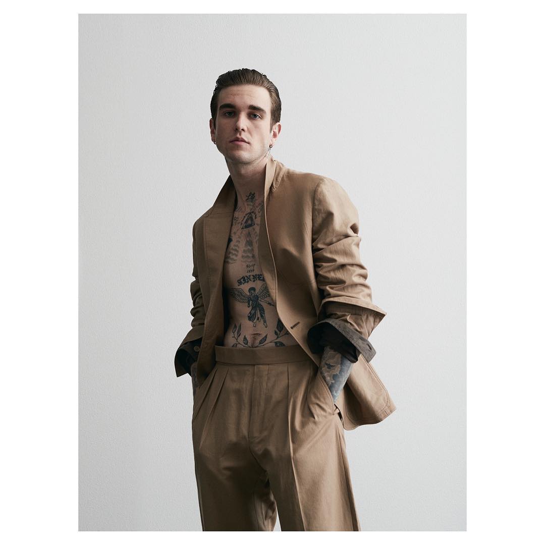 Man with chest tattoos wearing a camel suit jacket and trousers against a white seamless background