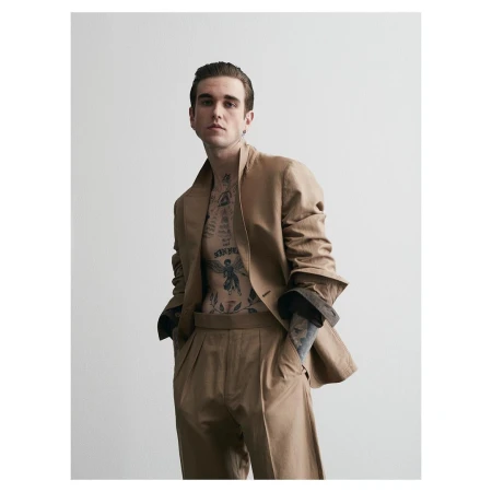 Man with chest tattoos wearing a camel suit jacket and trousers against a white seamless background