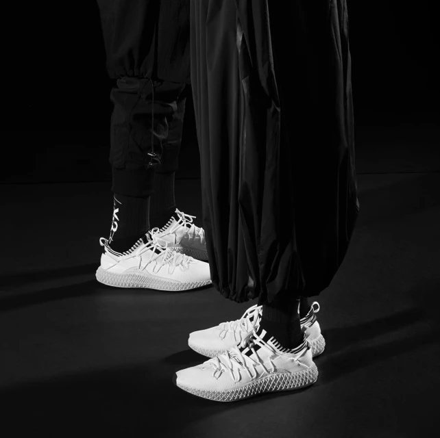 Two people wearing black athletic pants and white athletic shoes are standing indoors on a dark surface in a black and white image.