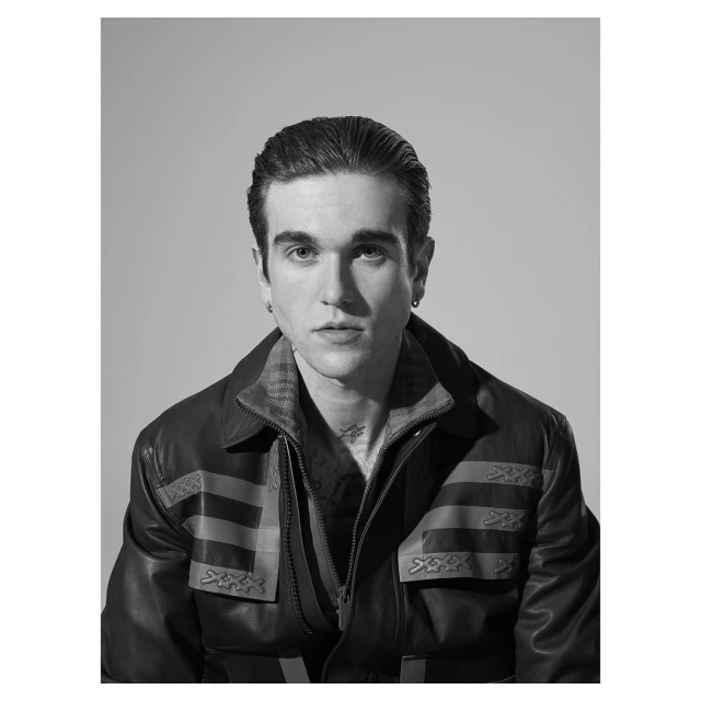 Black-and-white portrait of a young adult male with dark, slicked-back hair wearing a leather jacket in a studio setting with soft lighting.