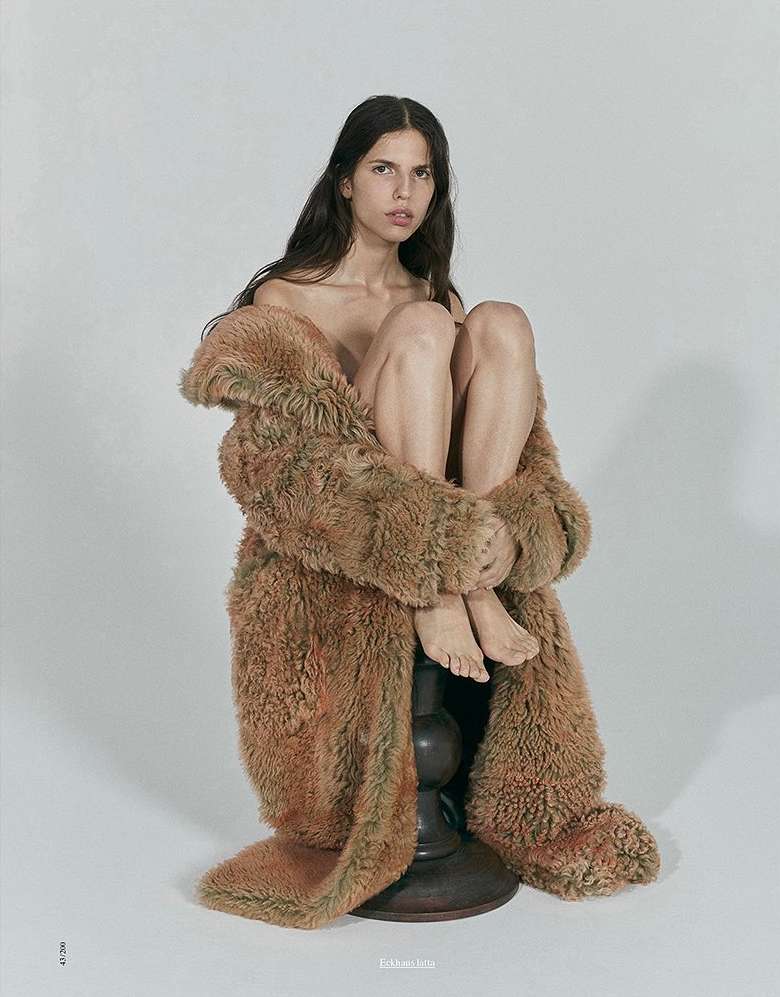 A woman with long brown hair sits on a dark wood base, enveloped in a furry, brown, floor-length coat against a plain white backdrop.