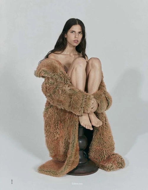 A woman with long brown hair sits on a dark wood base, enveloped in a furry, brown, floor-length coat against a plain white backdrop.