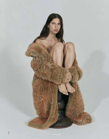 A woman with long brown hair sits on a dark wood base, enveloped in a furry, brown, floor-length coat against a plain white backdrop.