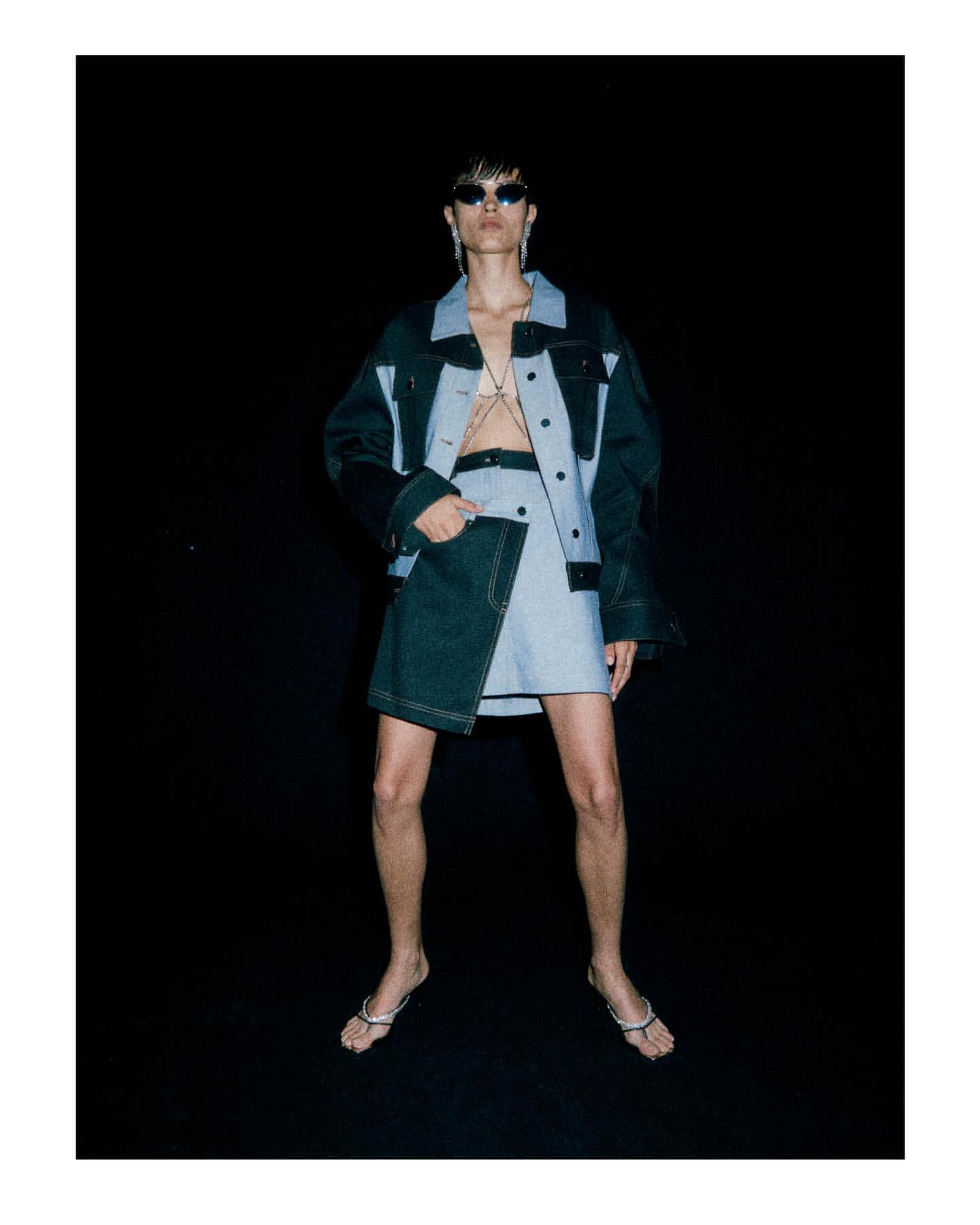 Fashion model wearing a two-tone denim jacket and mini skirt with silver jewelry poses in front of a black studio backdrop.