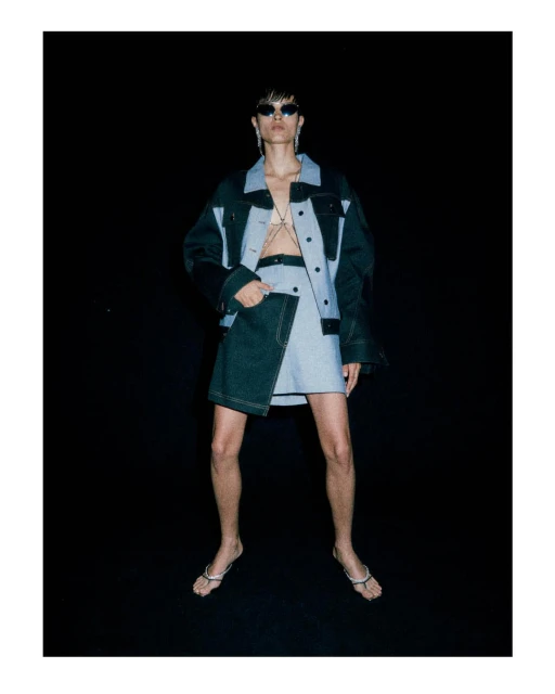 Fashion model wearing a two-tone denim jacket and mini skirt with silver jewelry poses in front of a black studio backdrop. 