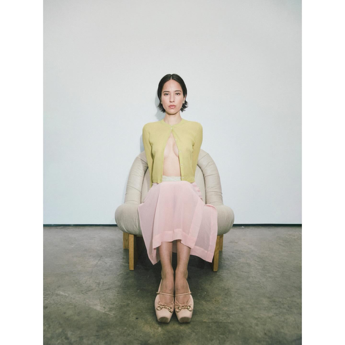 Young woman poses in a light-yellow cardigan, pink skirt, and ballet flats while seated in a beige armchair against a white background.