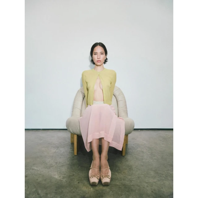 Young woman poses in a light-yellow cardigan, pink skirt, and ballet flats while seated in a beige armchair against a white background.