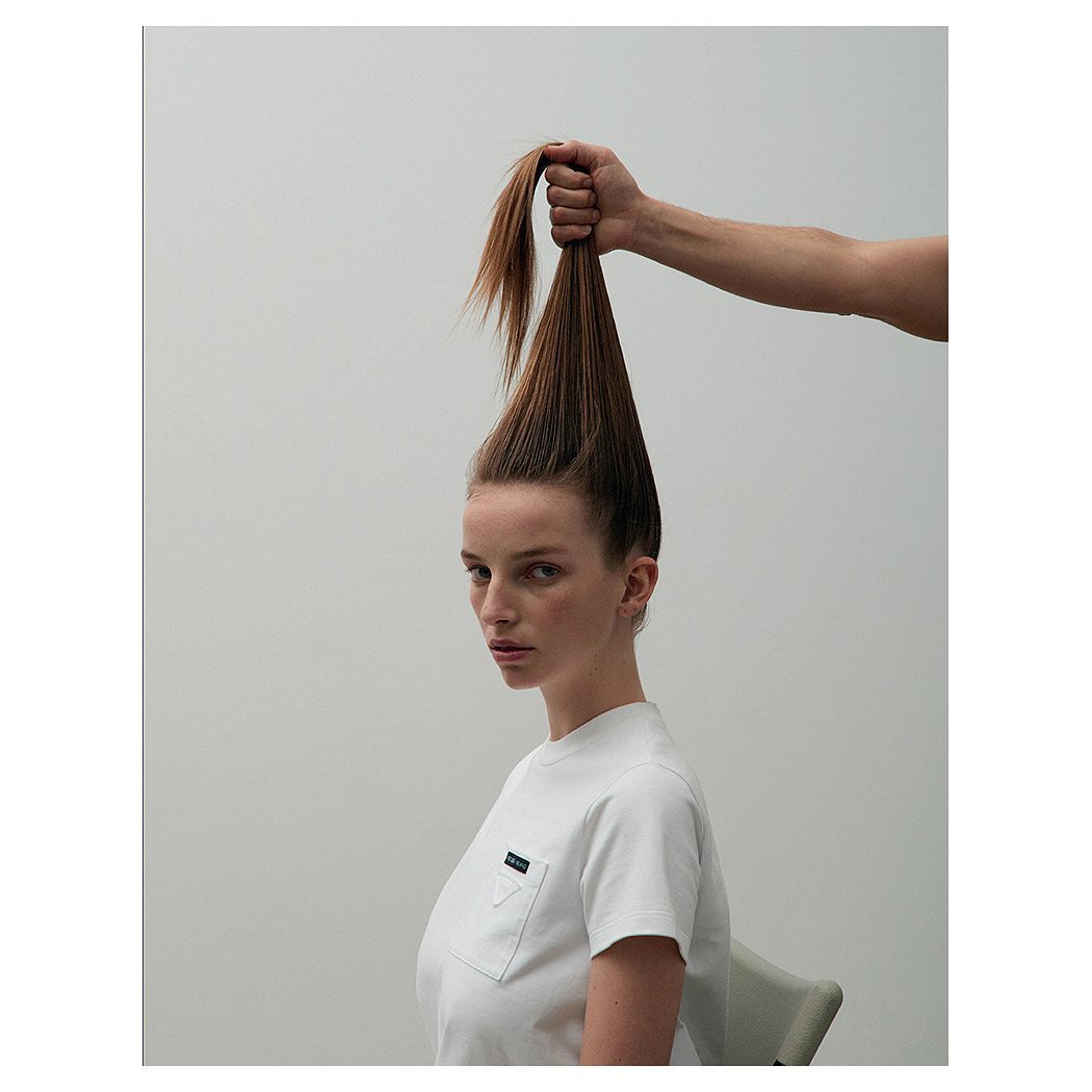 Eye-level studio shot of a young woman with ginger hair held high by a hand, wearing a white Prada t-shirt, set against a grey background.