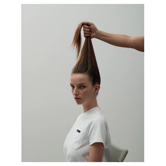 Eye-level studio shot of a young woman with ginger hair held high by a hand, wearing a white Prada t-shirt, set against a grey background.