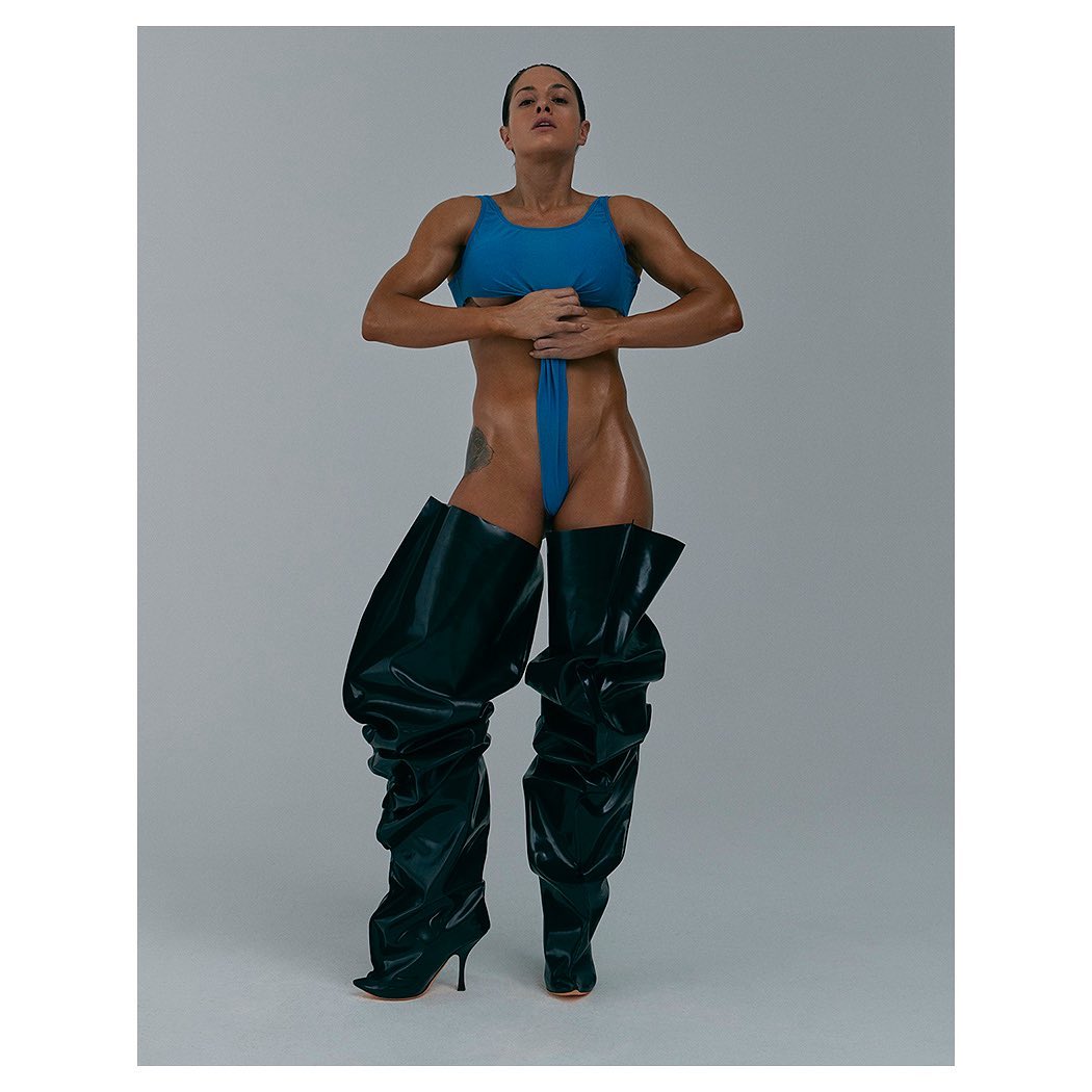 Muscular woman in blue swimsuit and shiny black, thigh-high heeled boots posing in a photo studio against a neutral grey backdrop.