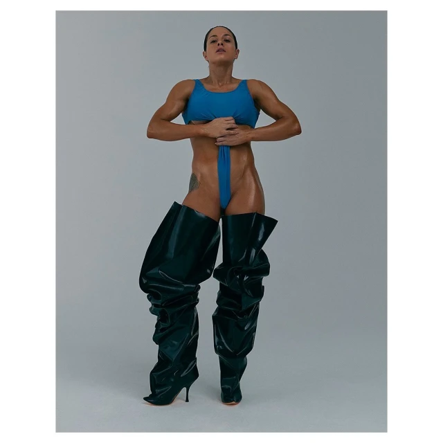 Muscular woman in blue swimsuit and shiny black, thigh-high heeled boots posing in a photo studio against a neutral grey backdrop.