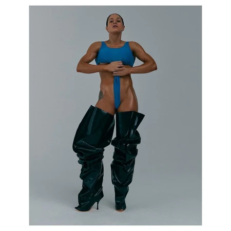 Muscular woman in blue swimsuit and shiny black, thigh-high heeled boots posing in a photo studio against a neutral grey backdrop.