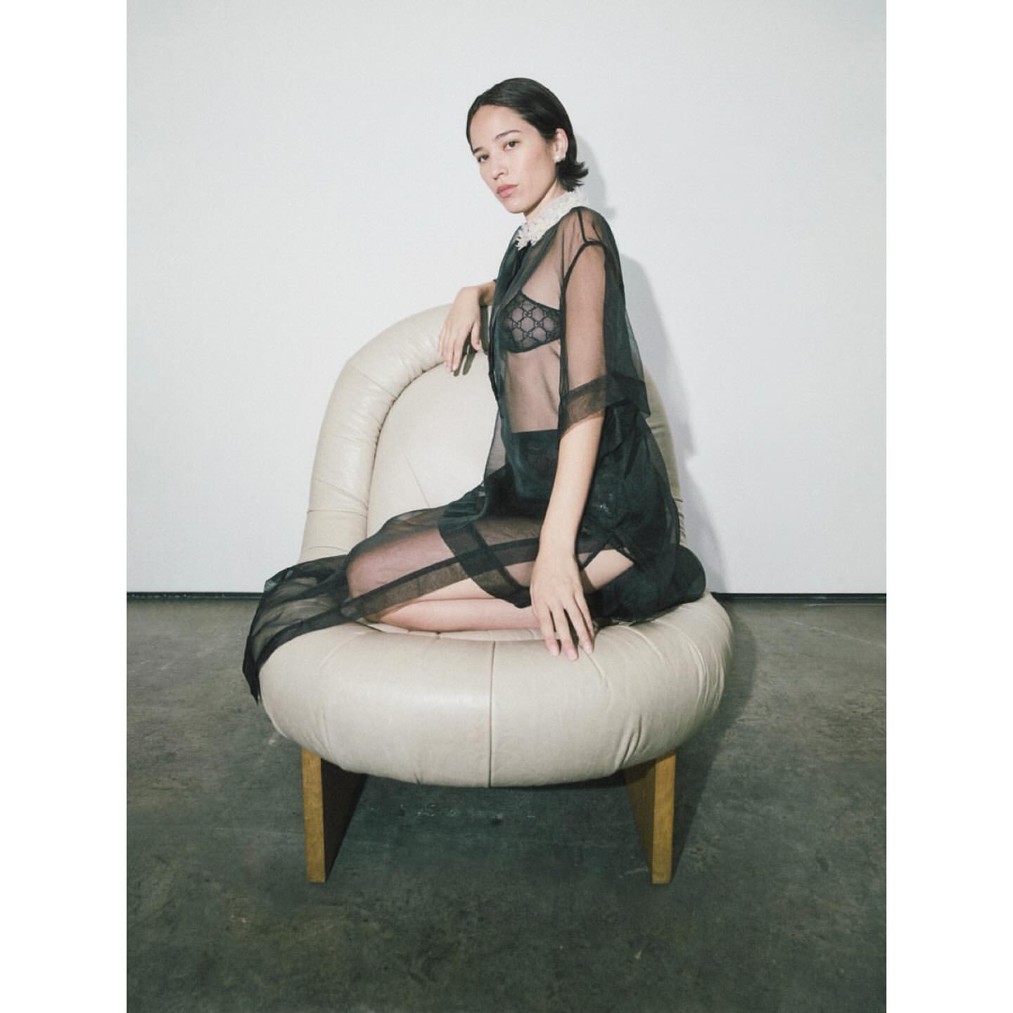 Young adult woman in semi-sheer black shirt and skirt seated on a round cream-colored chair in a minimalist indoor setting, fashion shot.