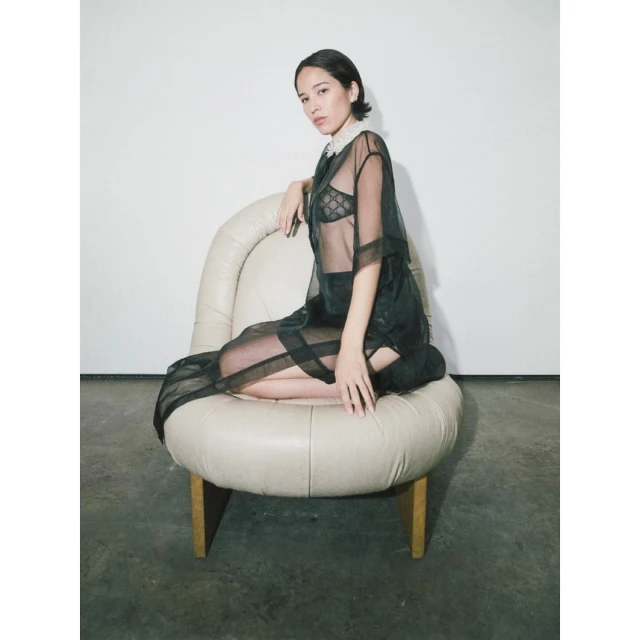 Young adult woman in semi-sheer black shirt and skirt seated on a round cream-colored chair in a minimalist indoor setting, fashion shot.