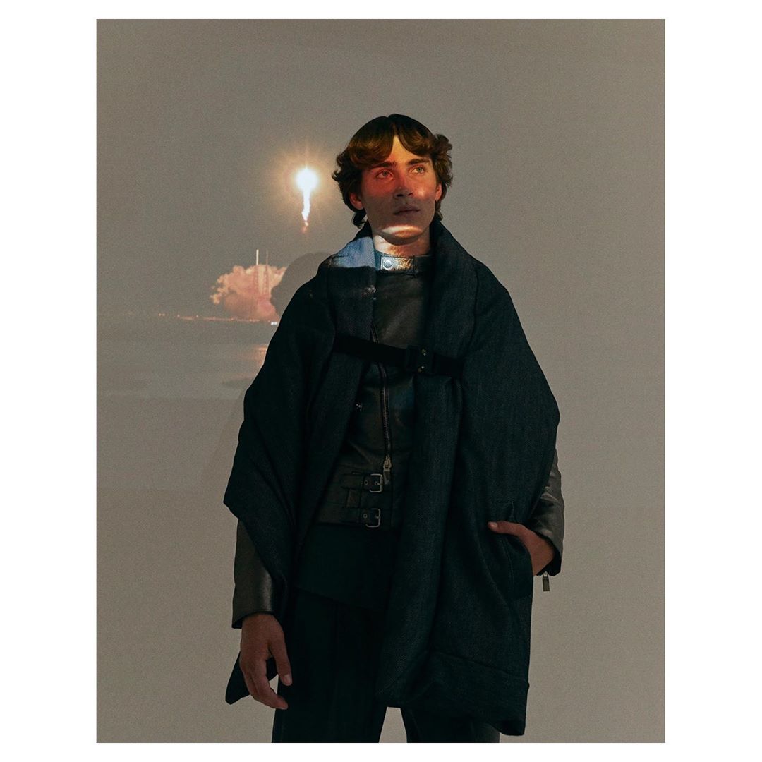 Young man poses in a stylish black outfit, with a space shuttle launch projected in the background, creating a modern, artistic portrait.