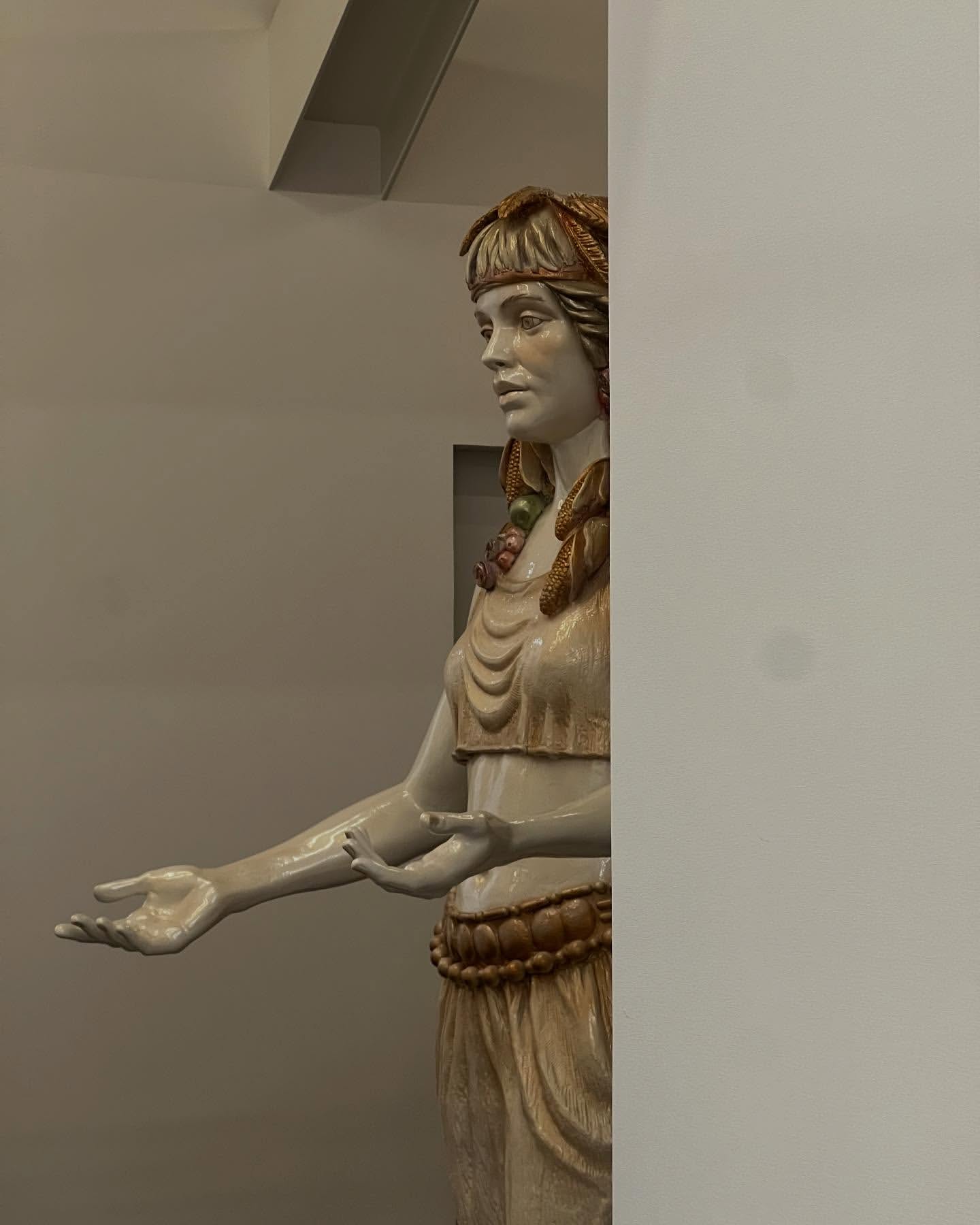 Statue of a fair-skinned woman wearing decorative headpiece, top, skirt, and belt against a neutral wall in an indoor setting.