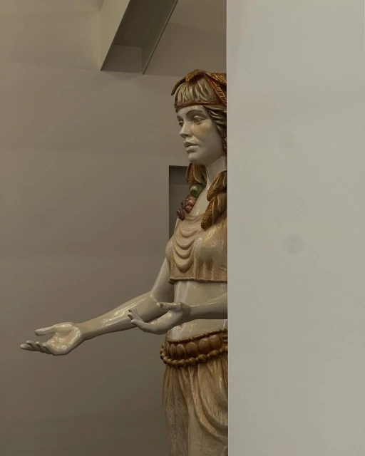 Statue of a fair-skinned woman wearing decorative headpiece, top, skirt, and belt against a neutral wall in an indoor setting.