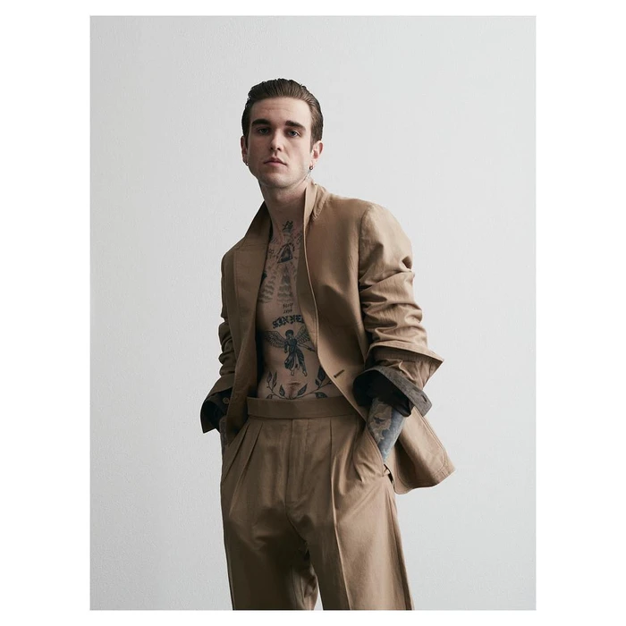 Man with chest tattoos wearing a camel suit jacket and trousers against a white seamless background