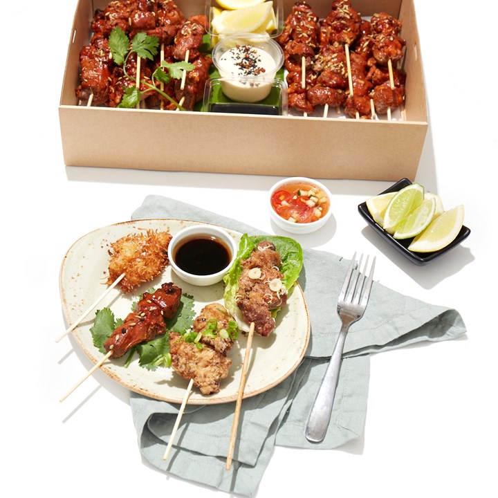 Skewered meat and seafood appetizers are arranged on a plate and in a take-out container, featuring vibrant garnishes and dipping sauces.